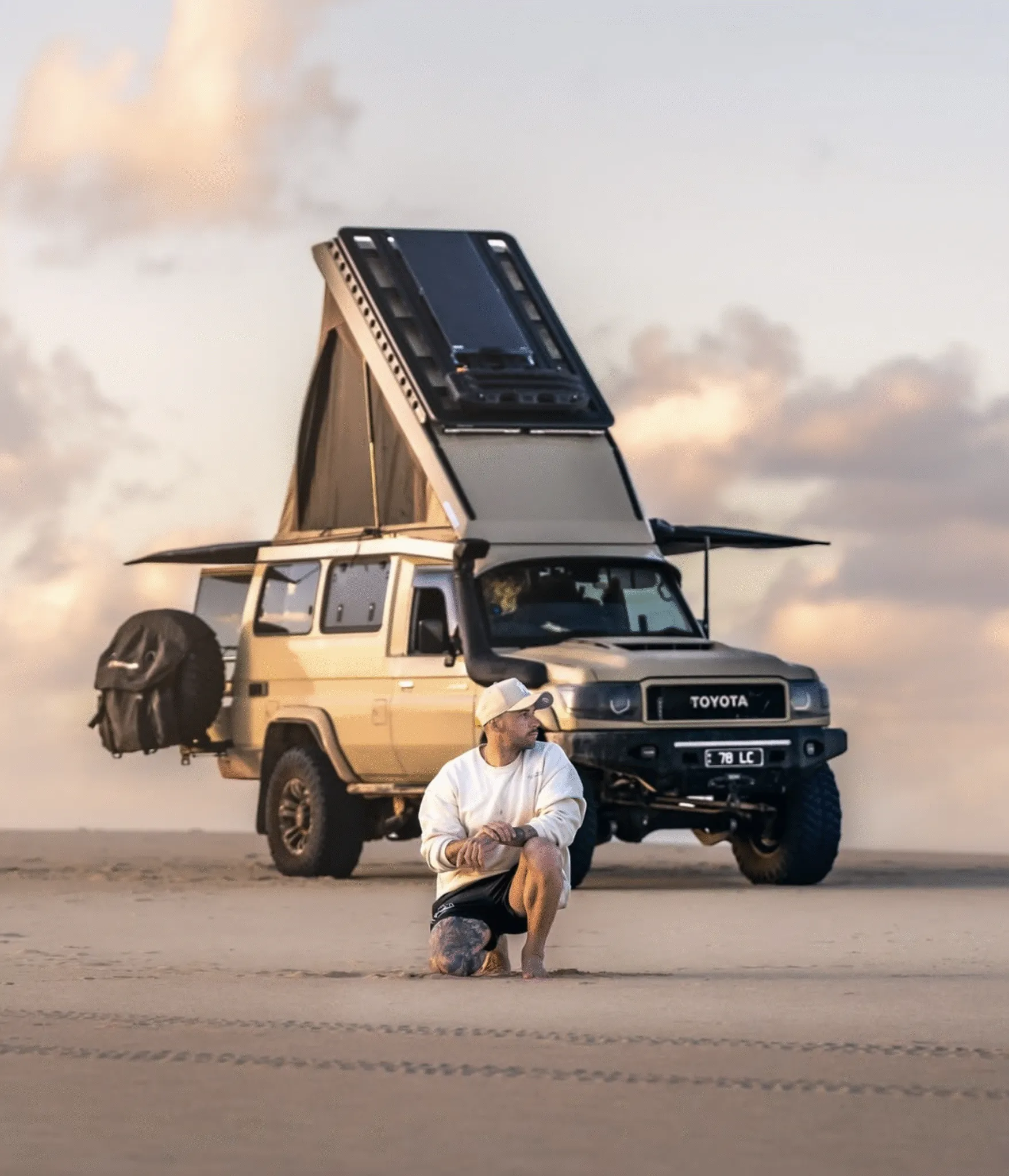 A man kneels on a sandy beach in front of a tan Toyota Land Cruiser Troop Carrier with a pop-top roof and solar panels.