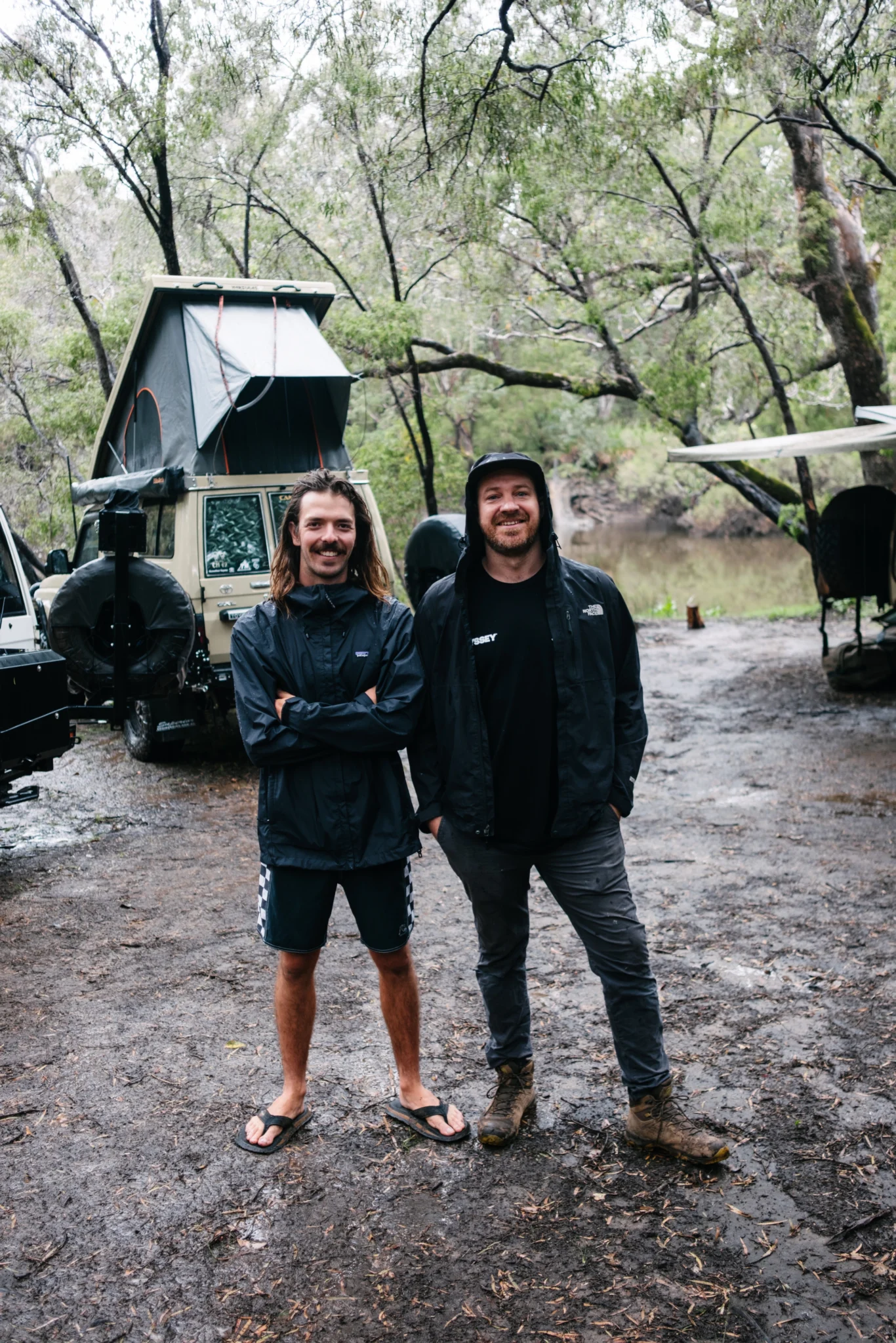 Two men stand in front of a Toyota Land Cruiser Troop Carrier with a pop-top roof in a muddy, wooded area.